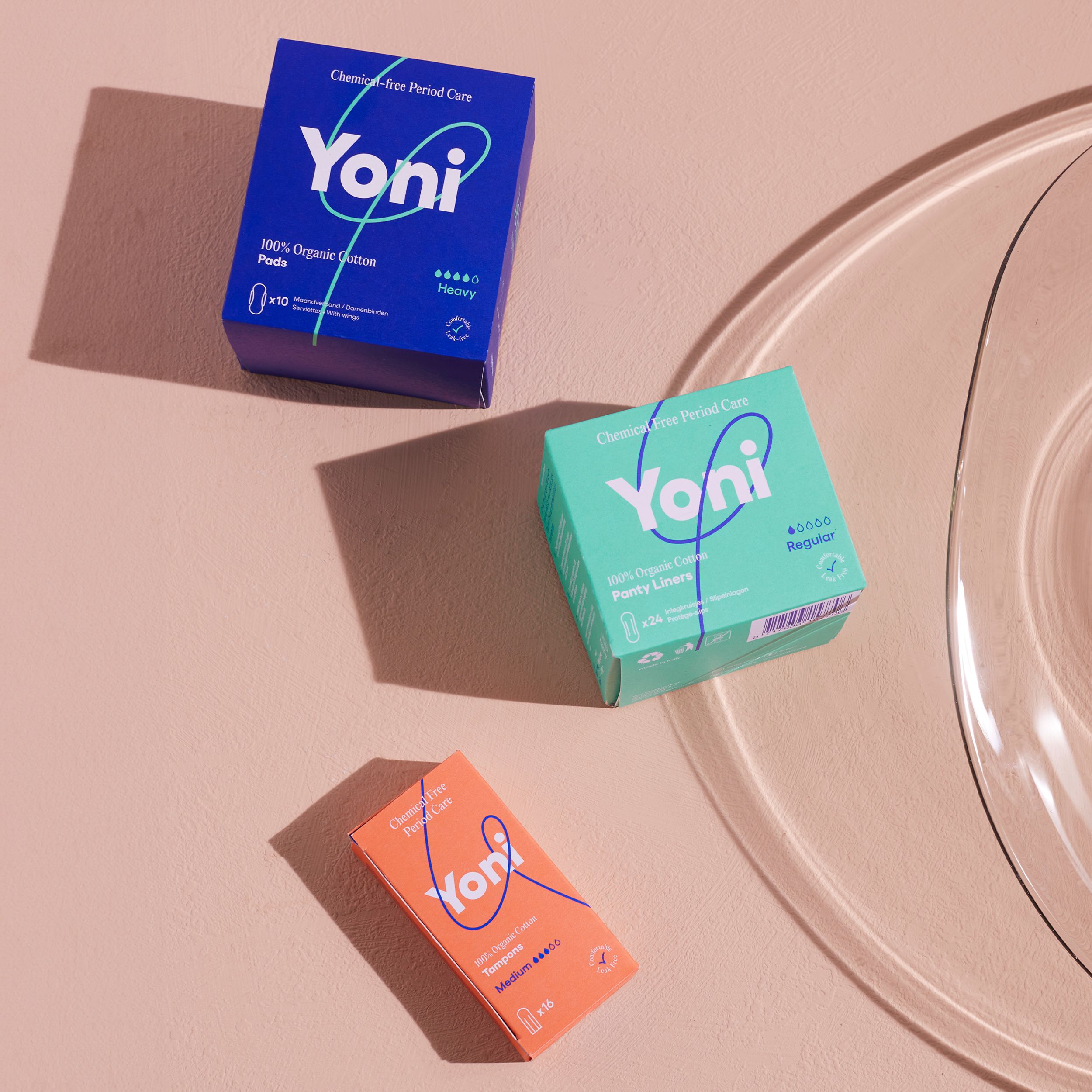 Trois boîtes de produits d'hygiène féminine. Logo Yoni. Bleu, vert et orange. Inscriptions : Pads, Pants Liner, Tampons.