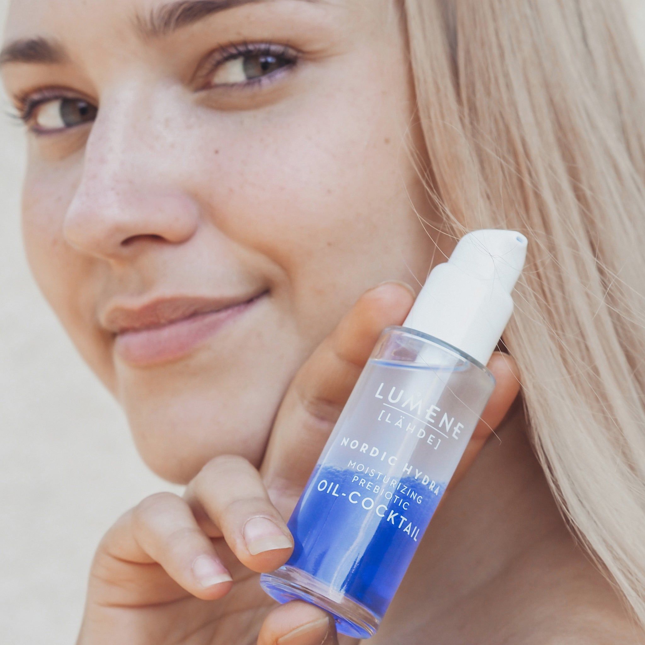 Femme tenant un flacon. Flacon avec pompe blanche. Liquide bleu et transparent visible. Nom du produit et marque.