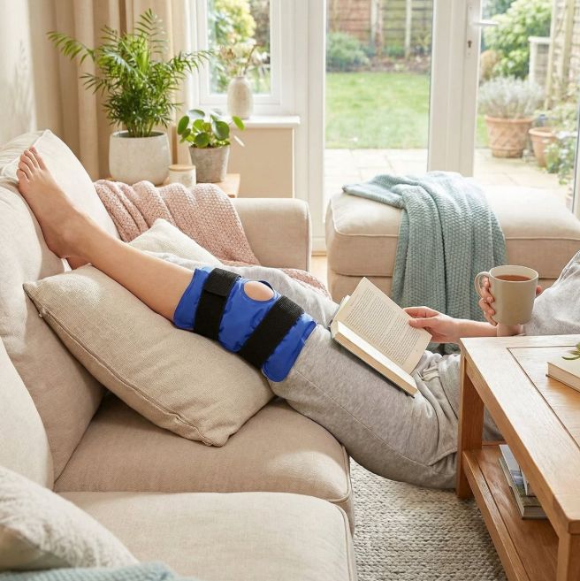 Persoon ligt op bank met kniebrace. Blauwe kniebrace met zwarte banden. Boek en kopje.
