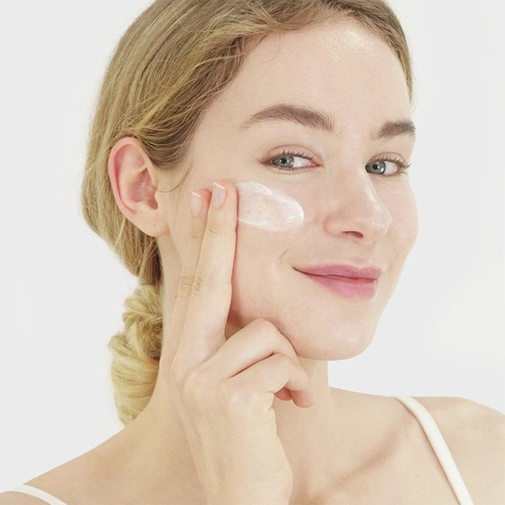 Vrouw brengt crème aan op haar gezicht. Ze glimlacht. Crème op vinger en huid. Lichte huid, blond haar.