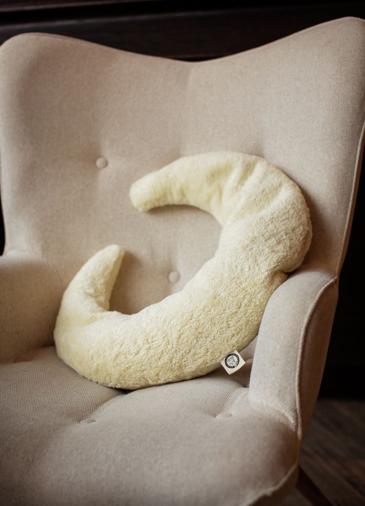 Coussin crème en forme de croissant sur un fauteuil beige. Le coussin est en matière moelleuse.
