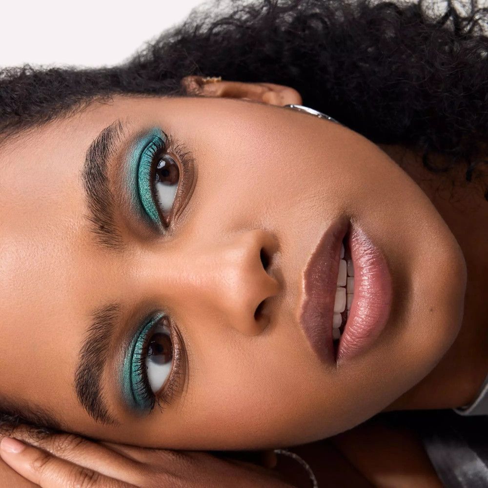 Gros plan d'un visage avec fard à paupières vert. Peau foncée, yeux bruns, bouche ouverte.