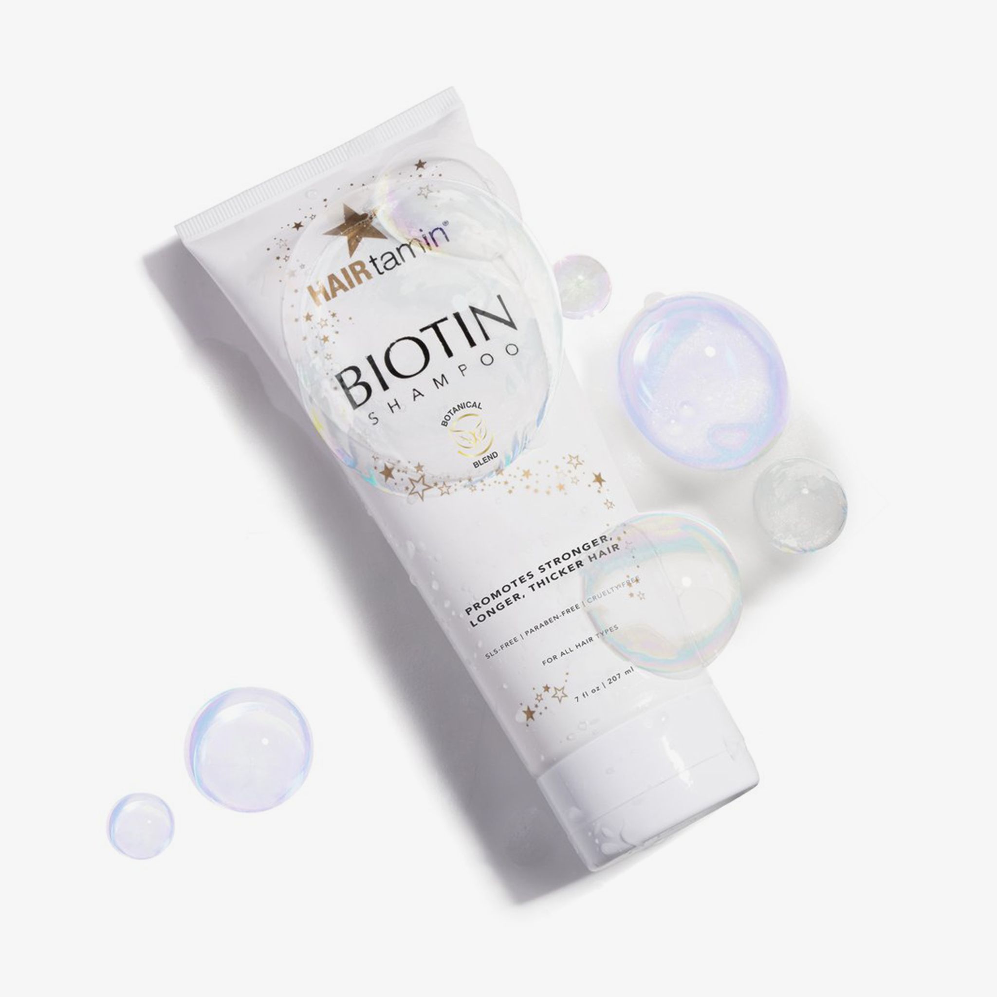 Tube blanc "HAIRtamin Biotin Shampoo" avec bulles de savon. Étoiles et texte "Botanical Blend". Texte : "Supports stronger, longer, thicker hair".