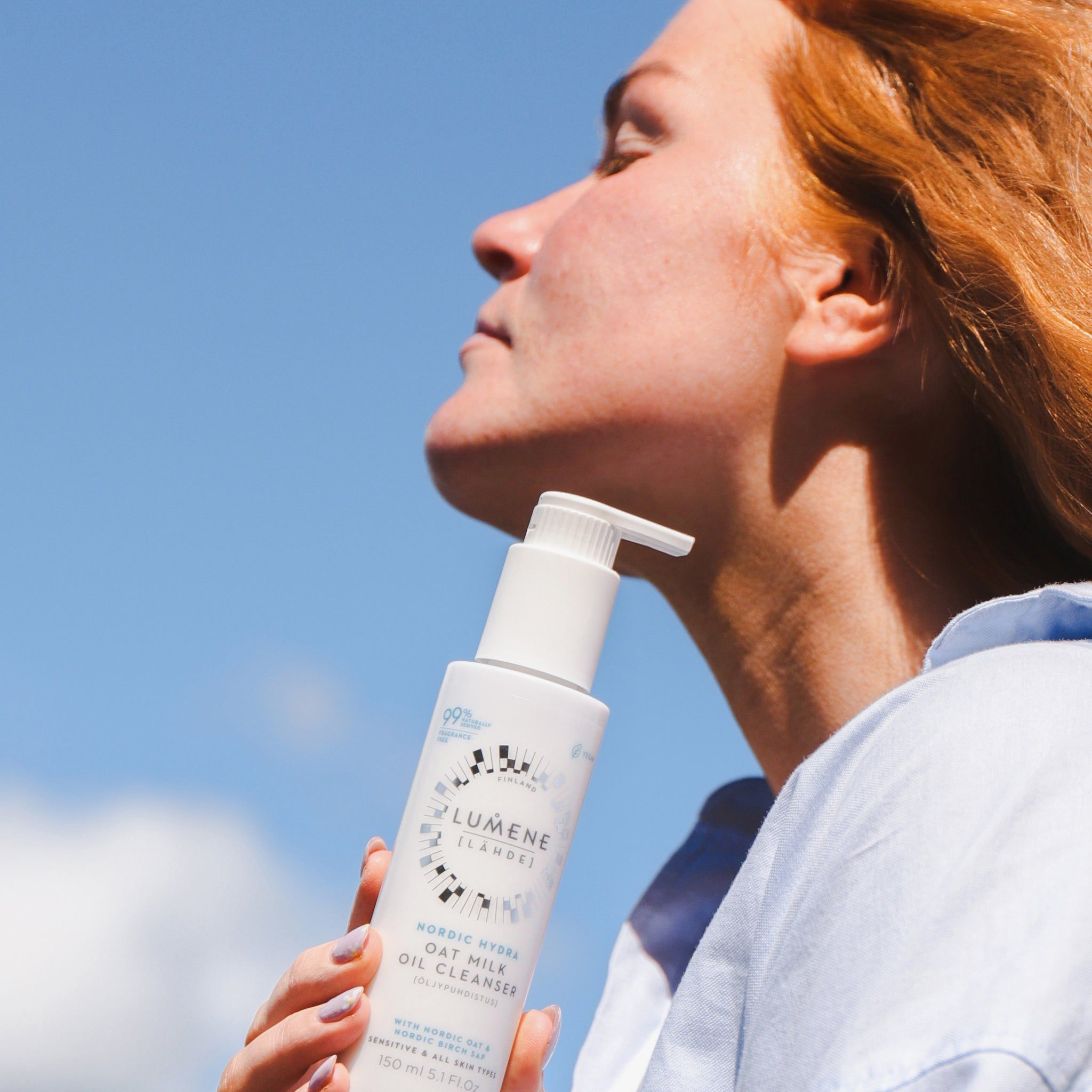 Vrouw houdt een witte fles met pomp vast. Opschrift: Lumene Nordic Hydra Oat Milk Oil Cleanser. 99% natuurlijk. Blauwe hemel.