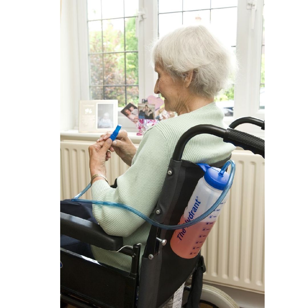 Oudere vrouw in rolstoel, drinkt uit een drinkfles met rietje. De fles is aan de rolstoel bevestigd.