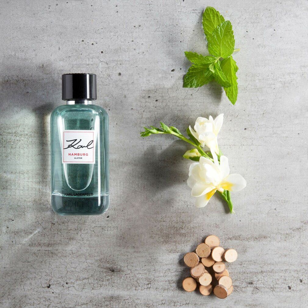 Karl Lagerfeld Hamburg Alster E.d.T. Nat. Spray. Flacon avec bouchon noir, à côté de fleurs et de morceaux de bois.
