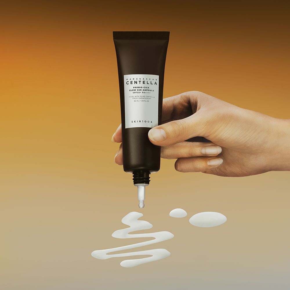 Main tenant un tube. Le produit coule. Texte: MADAGASCAR CENTELLA, PROBIO-CICA GLOW SUN AMPULLE SPF50+ PA++++. Gouttes jaunes.