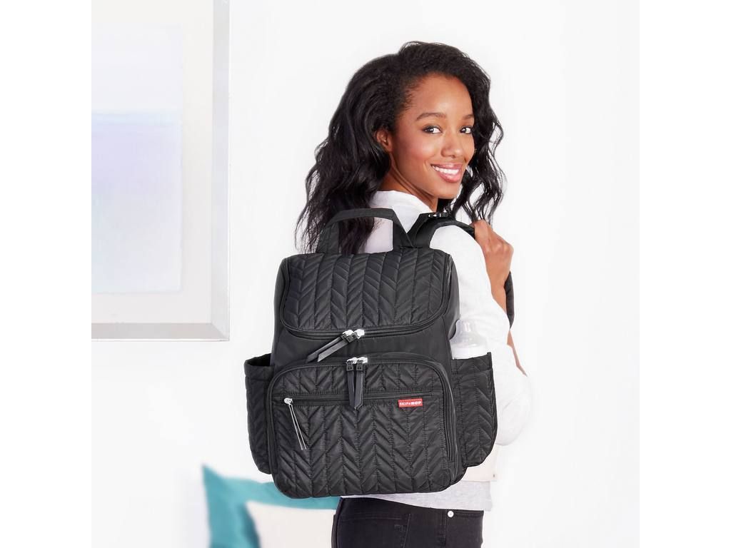 Zwarte luiertas op de rug van een vrouw. Zigzagpatroon, meerdere vakken en ritsen. Rode merknaam.