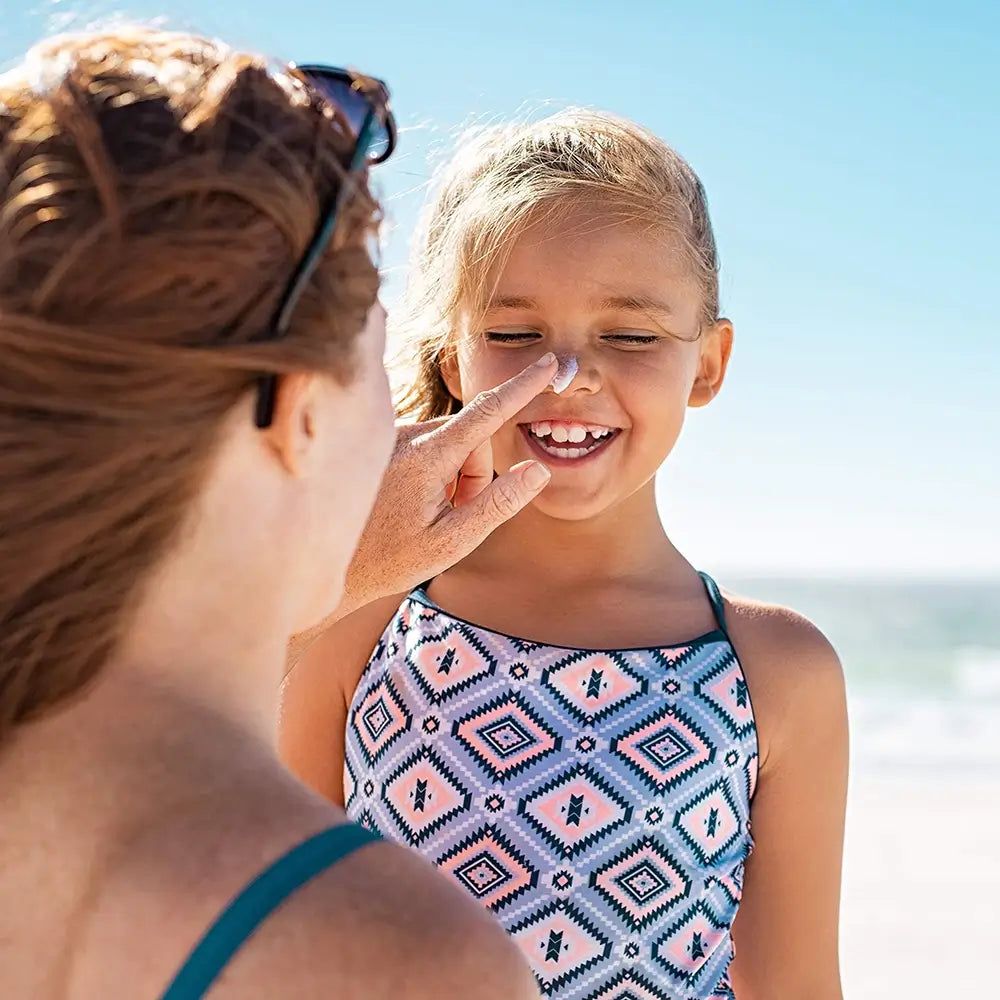 Une mère applique de la crème solaire sur le nez de son enfant. L'enfant porte un maillot de bain à la plage.