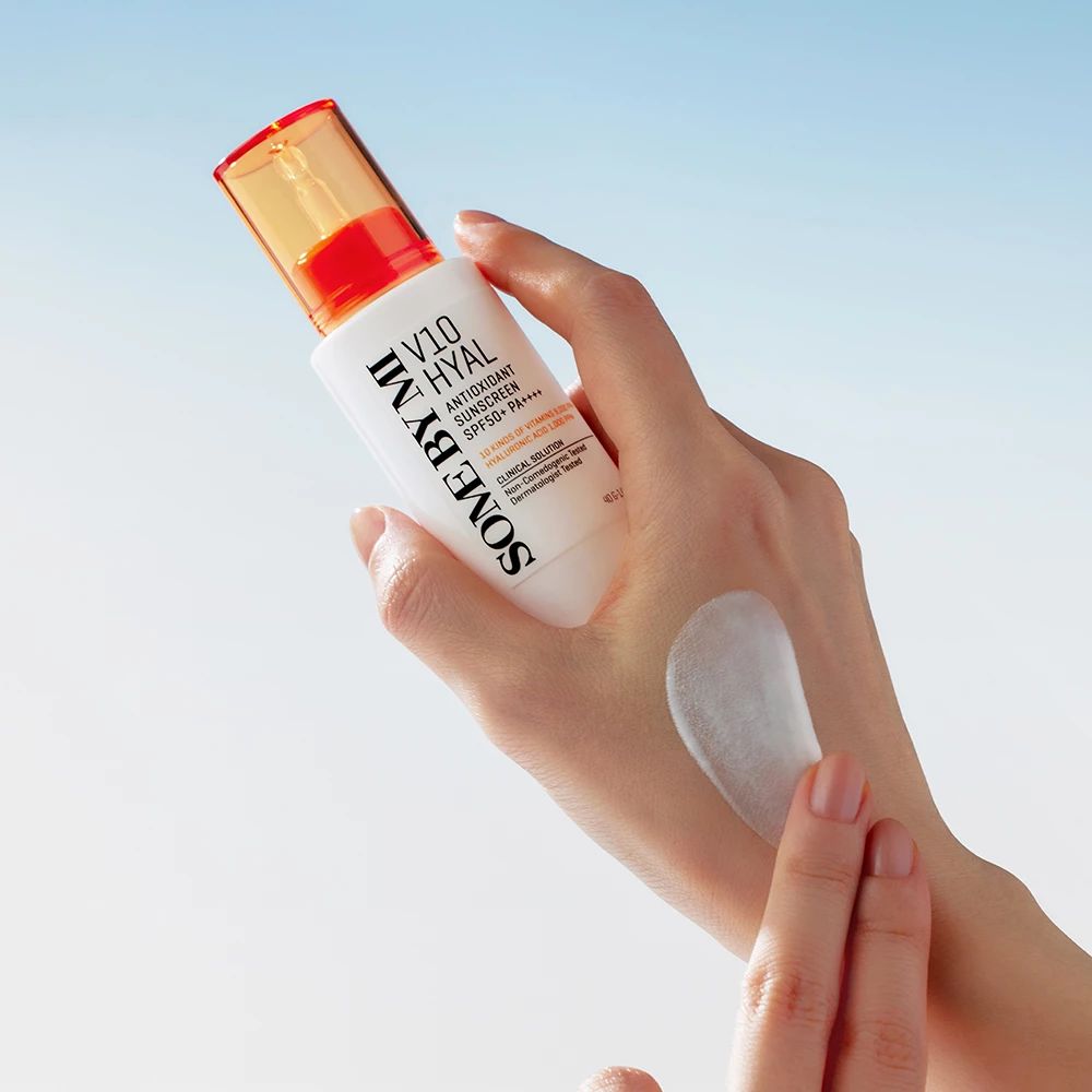 Flacon blanc avec bouchon orange. Produit appliqué sur la main. Texte : SOME BY MI, V10 HYAL Antioxidant Sunscreen SPF50+.