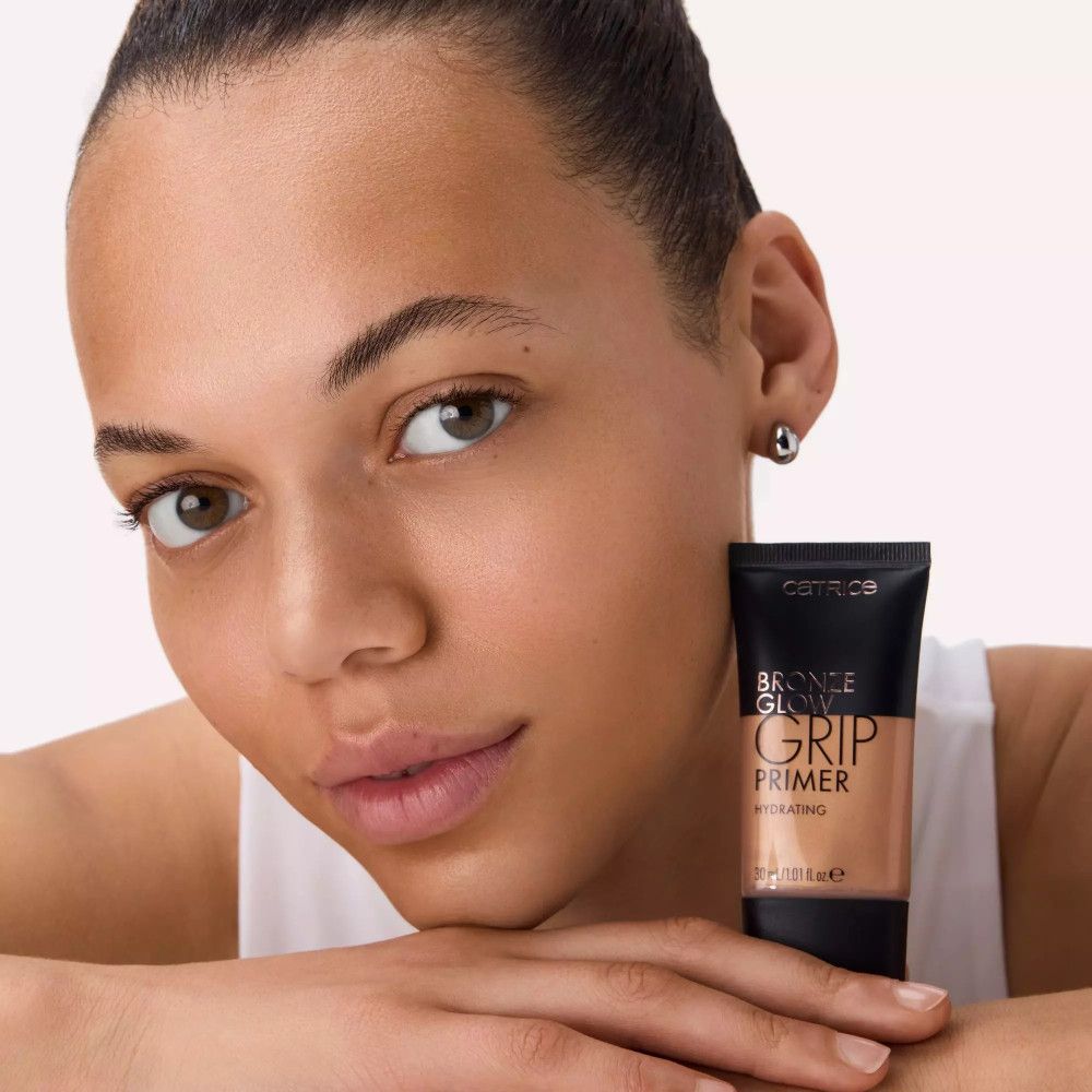 Femme tenant un tube Bronze Glow Grip Primer. Produit à côté du visage. Tube noir avec nom du produit.