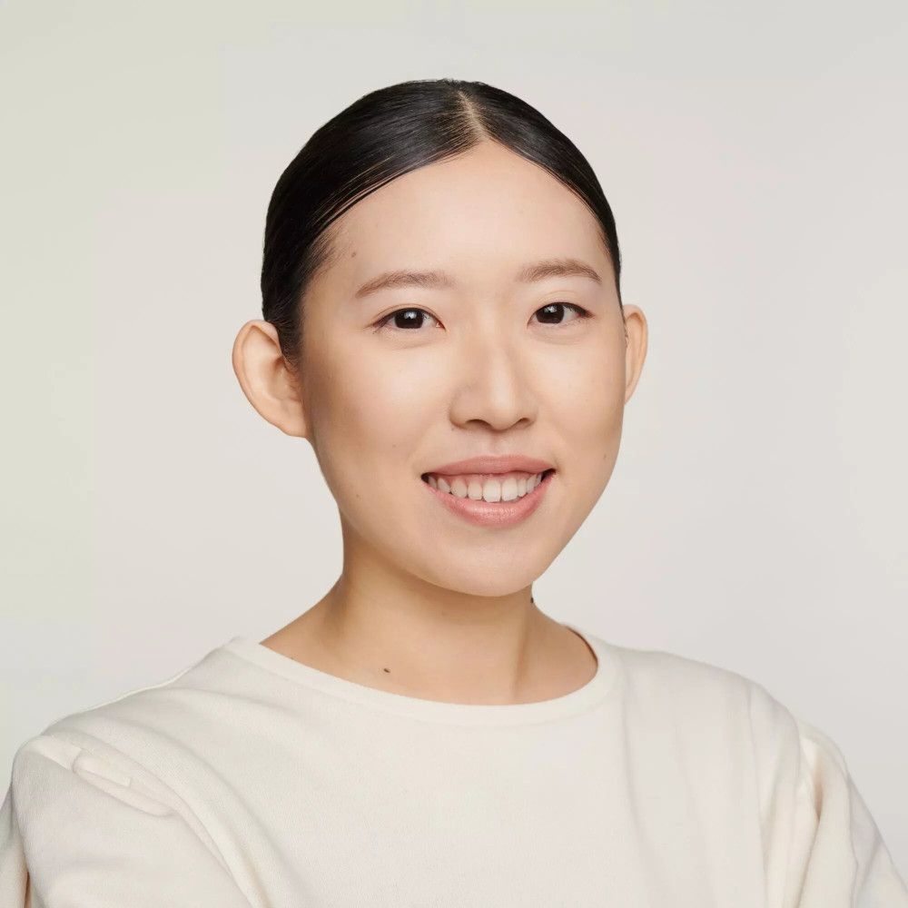 Portrait d'une femme souriante aux cheveux foncés. Elle porte un haut crème. Fond clair.