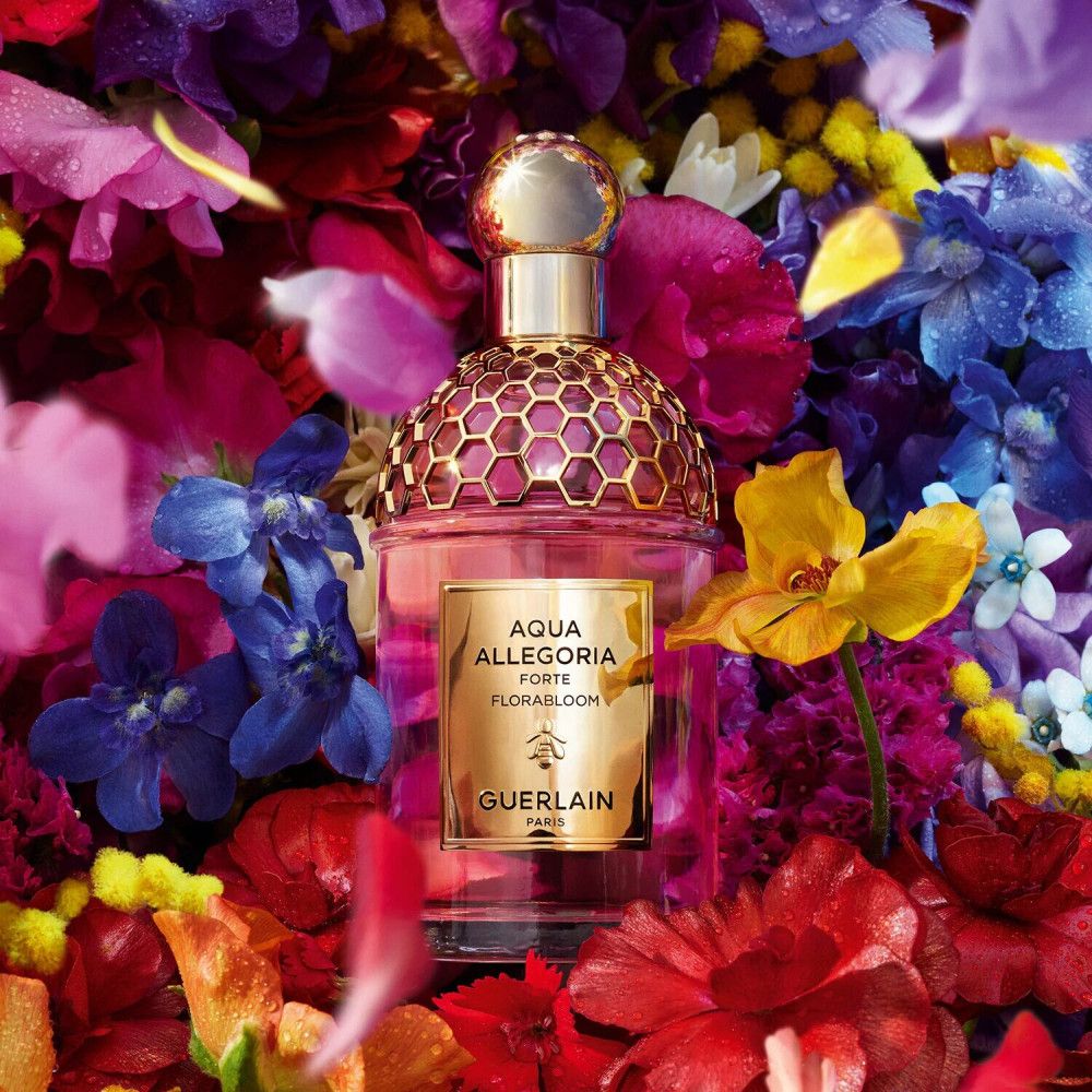 Roze parfumfles met gouden dop, omringd door kleurrijke bloemen. Opschrift: Aqua Allegoria Forte Flora Bloom, Guerlain.