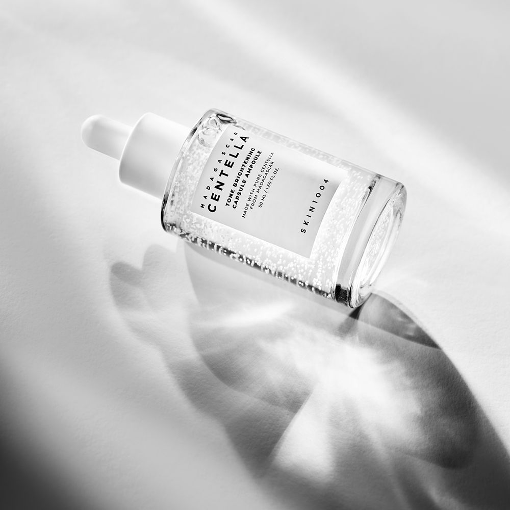Schuin liggende glazen fles met witte dop. Bevat vloeistof met kleine deeltjes. Tekst: MADAGASCAR CENTELLA, Tone Brightening Capsule Ampoule.