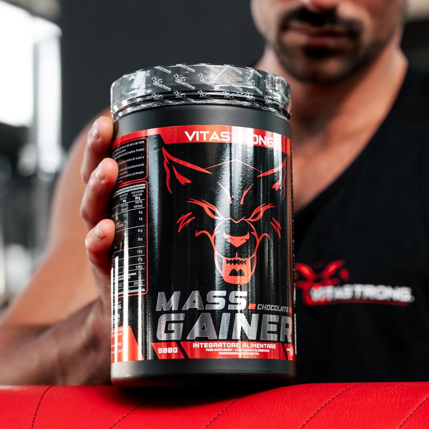Vitastrong Mass Gainer-container. Zwarte container met rood label. De container wordt door een persoon vastgehouden.