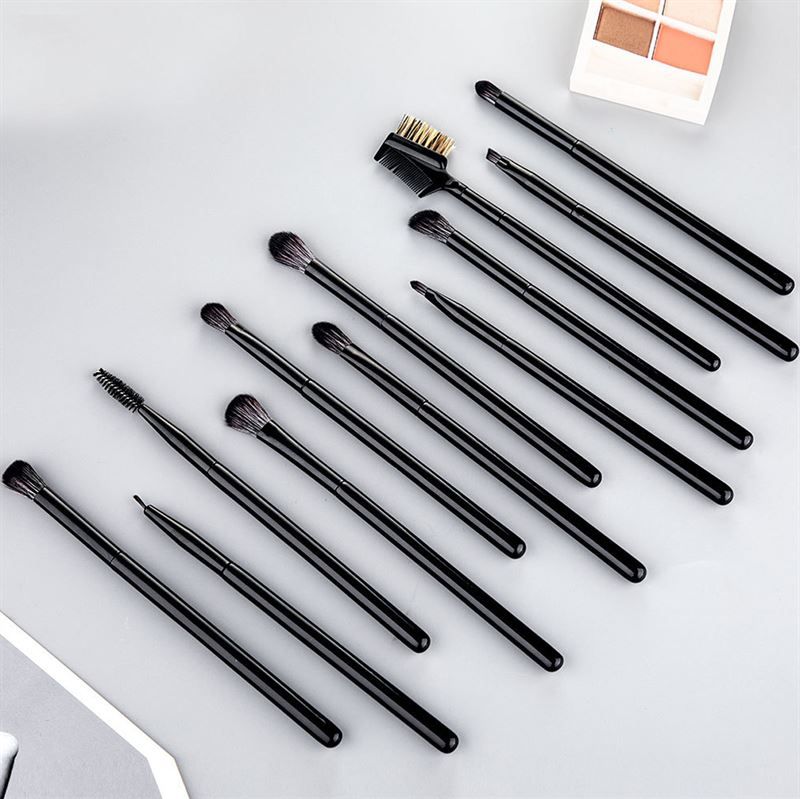 Douze pinceaux noirs pour fard à paupières et sourcils, posés sur une surface grise, avec un set de fard à paupières en arrière-plan.