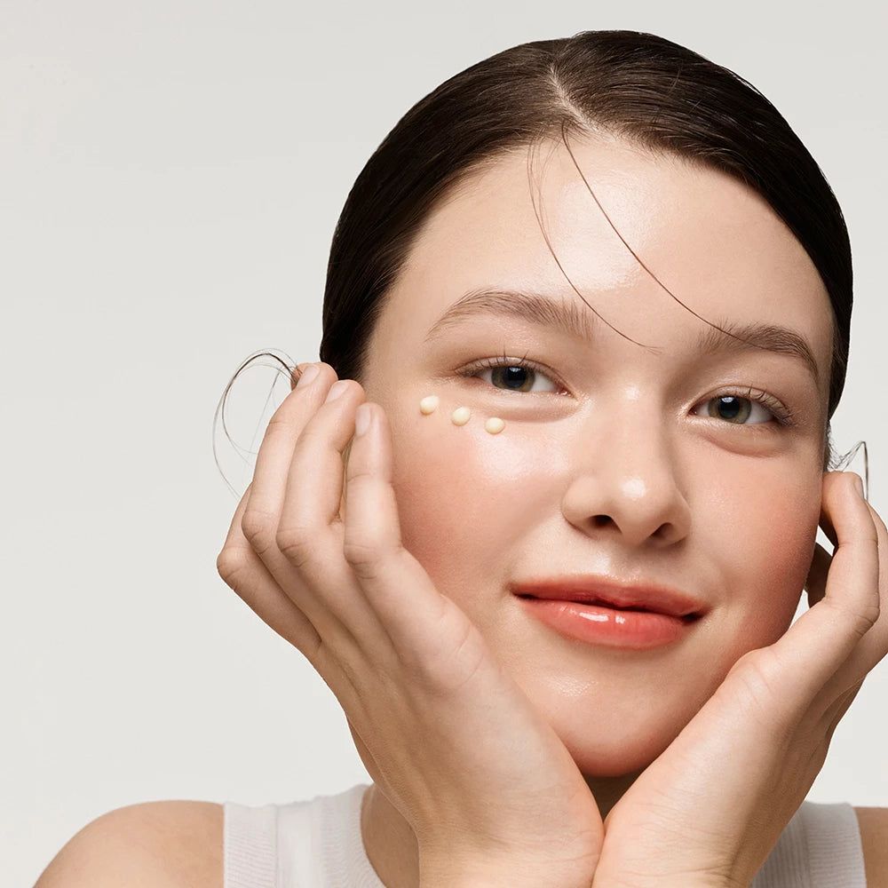Portrait d'une femme avec des points de crème sous les yeux. Elle sourit. Mains sur le visage.