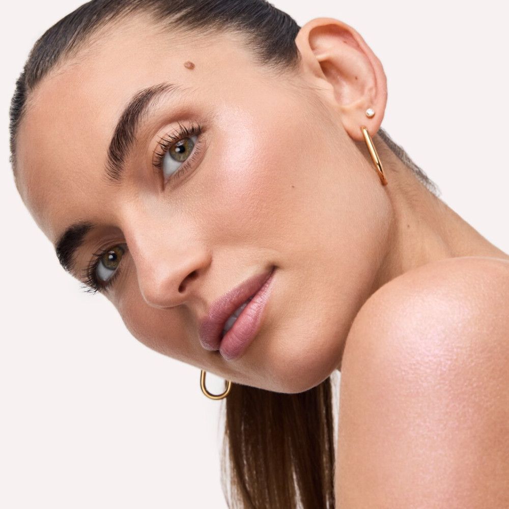Gros plan d'une femme avec de l'highlighter sur la peau. Elle porte des boucles d'oreilles dorées. Elle regarde la caméra.