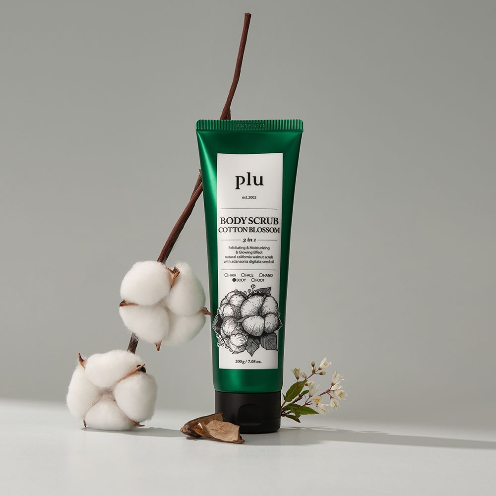 Tube vert avec étiquette blanche. Inscription : PLU, BODY SCRUB COTTON BLOSSOM. Illustration : fleurs de coton. Branches et fleurs de coton. Fond gris.