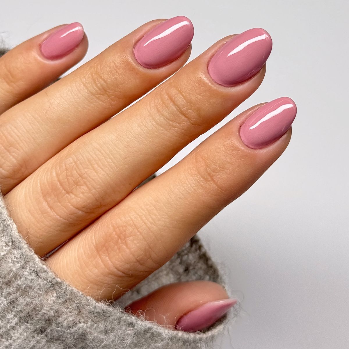 Main avec ongles roses ovales. Les ongles sont vernis brillants. La main porte un pull gris.