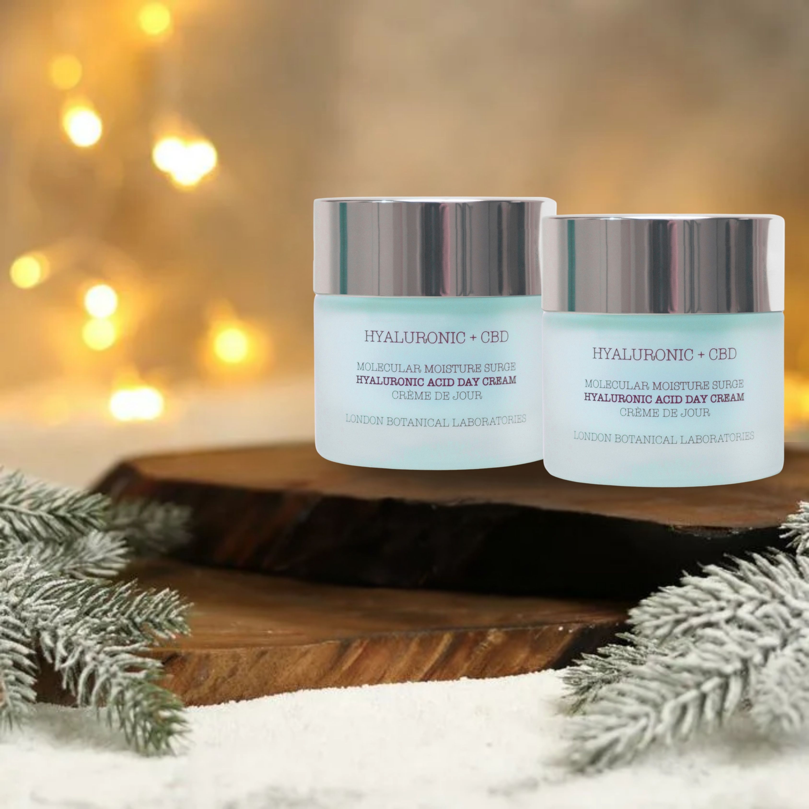 Twee potten dagcrème met tekst: Hyaluronzuur + CBD. Op een houten plank, met dennentakken en bokeh-effect op de achtergrond.