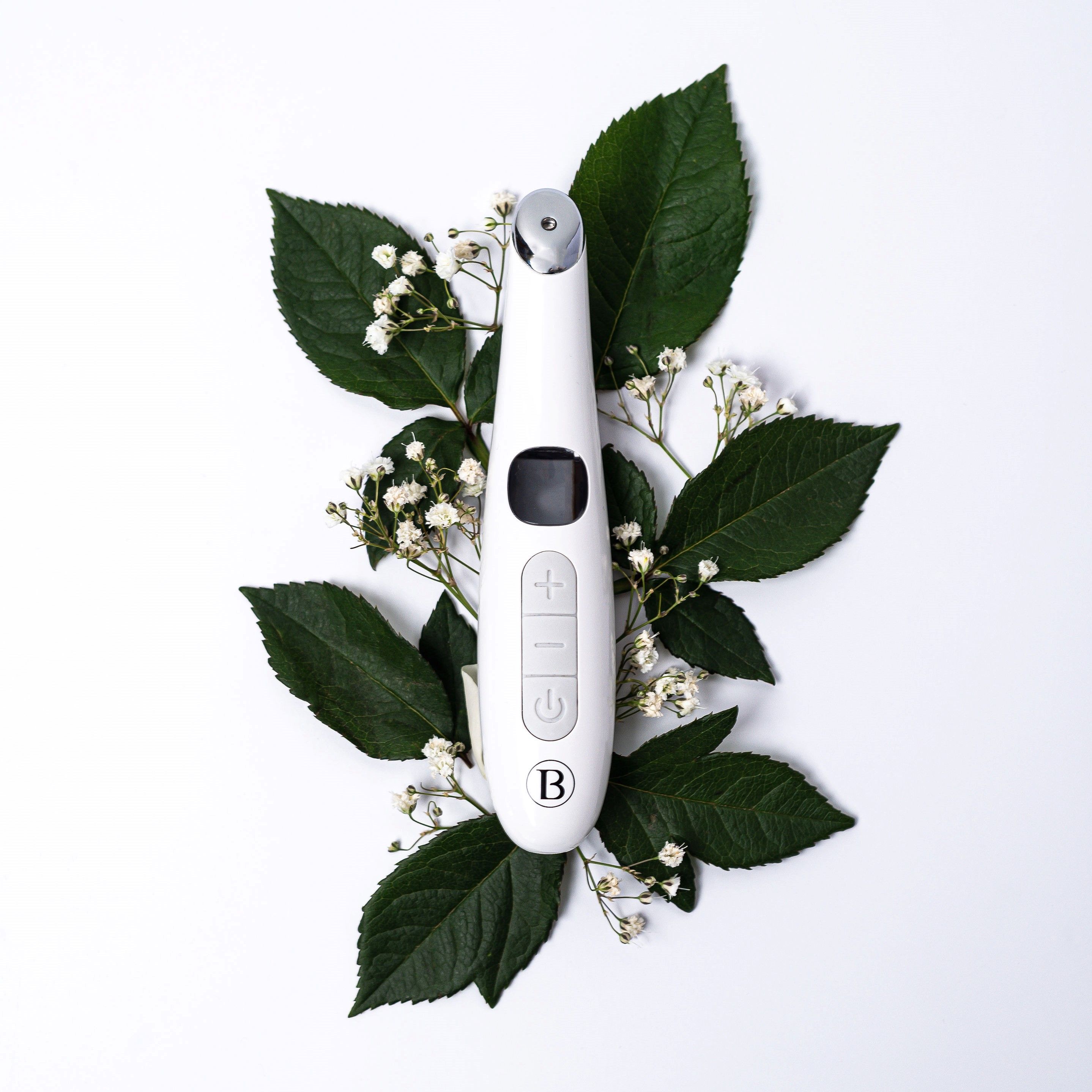 Appareil blanc avec écran, boutons et pointe rouge. Entouré de feuilles vertes et de fleurs blanches.