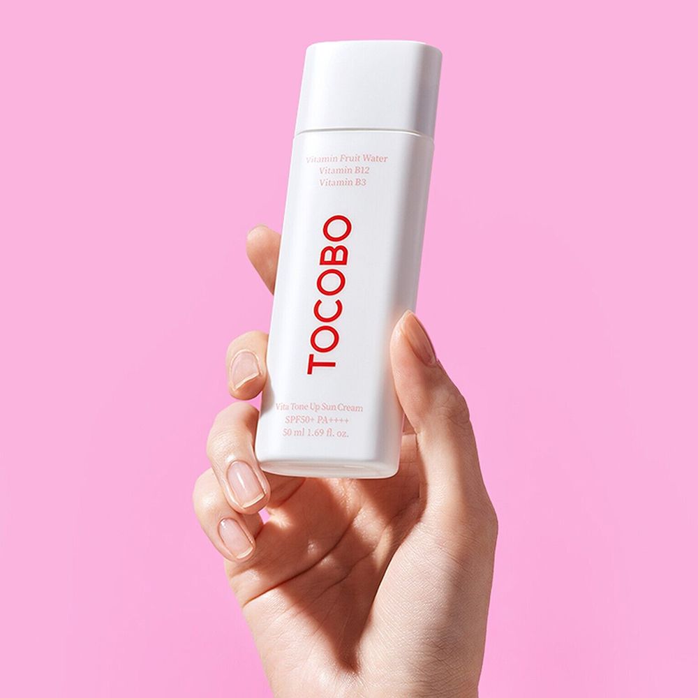 Witte tube met rood TOCBO-logo, vastgehouden door een hand. Bevat Vitamin Fruit Water, B12 en B3. SPF50+ PA++++.