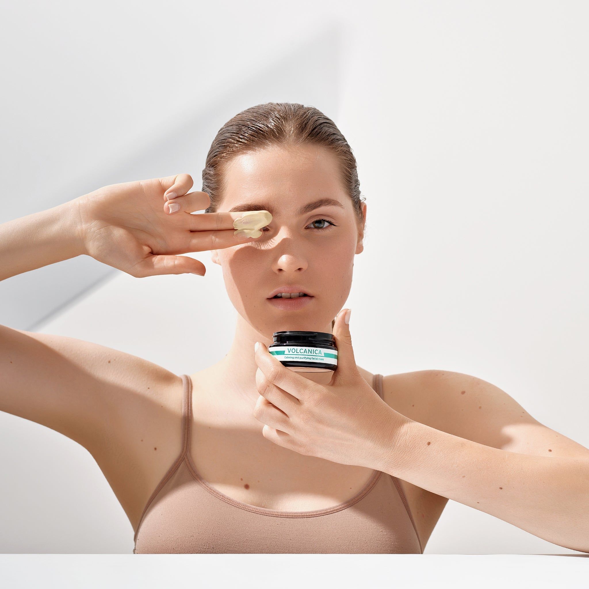 Vrouw houdt potje vast en brengt masker aan. Product: VOLCANICA. Lichte huid, hemdje.