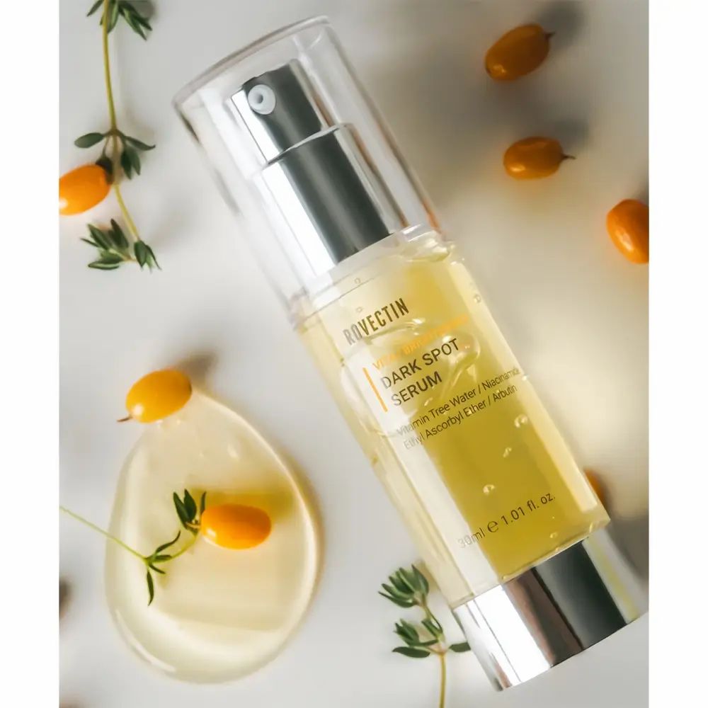 Serumfles met pomp, omringd door kumquats en takken. Opschrift: ROVECTIN Vita Dark Spot Serum. Bevat vitamine water, etc.