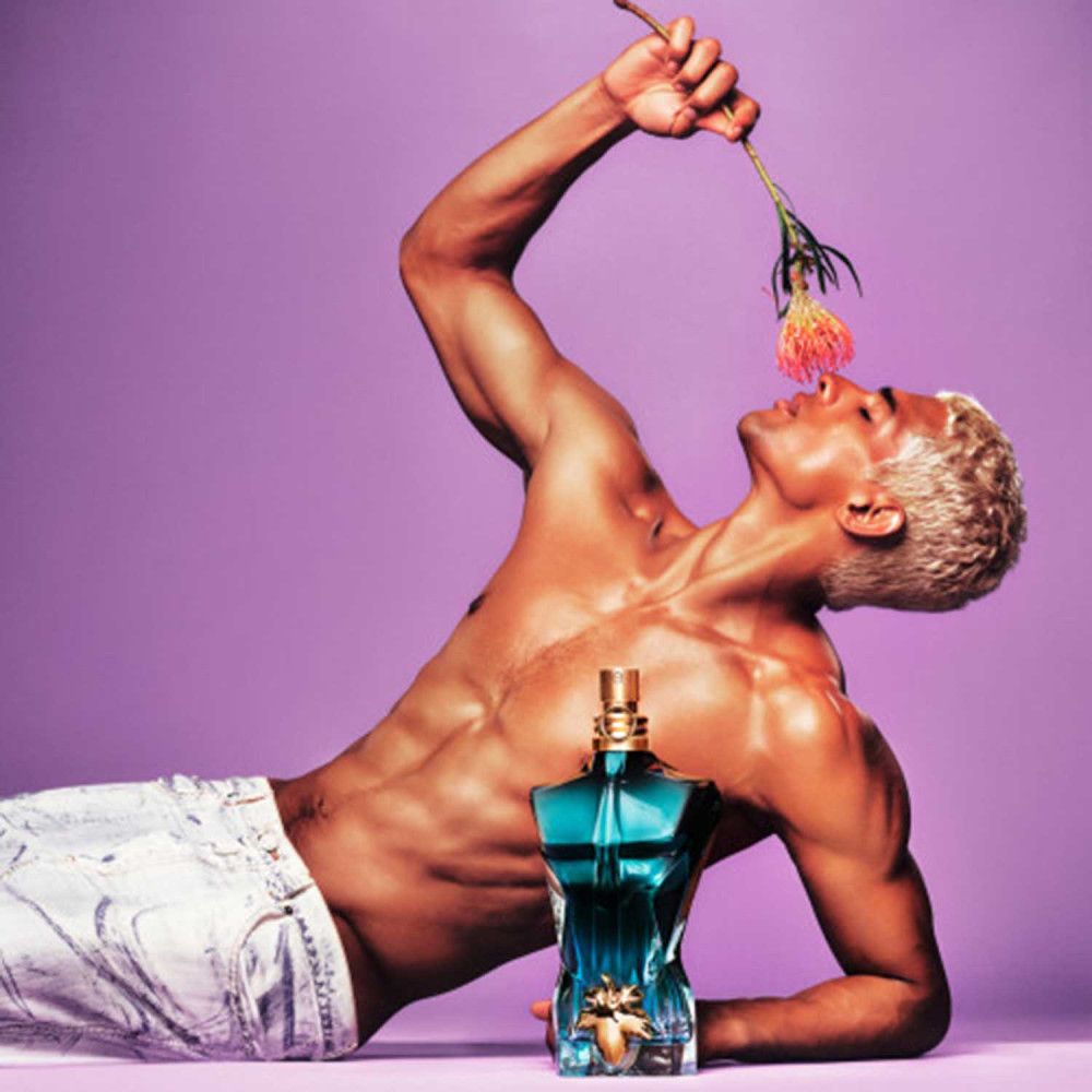 Homme blond tenant une fleur. Flacon Jean Paul Gaultier "Le Beau" en forme de torse. Sur fond violet.