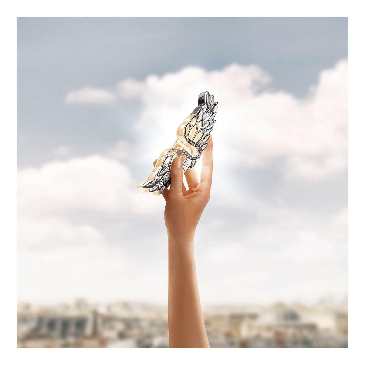 Main tenant un pendentif en forme d'ailes argentées devant un ciel bleu nuageux.