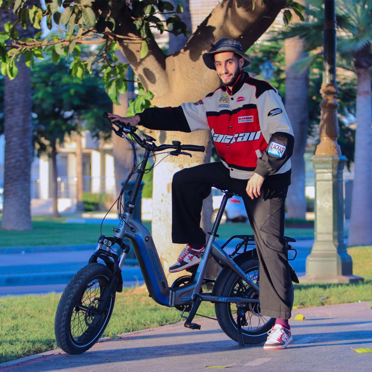 Persoon zit op een elektrische fiets. De man draagt een jas en een hoed. De fiets is grijs en blauw. De achtergrond is een straat.