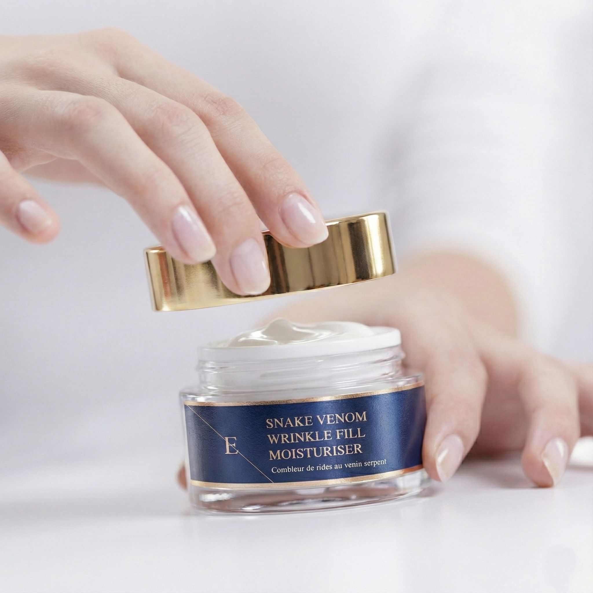 Mains ouvrant un pot de crème. Inscription: Snake Venom Wrinkle Fill Moisturiser. Couvercle doré.