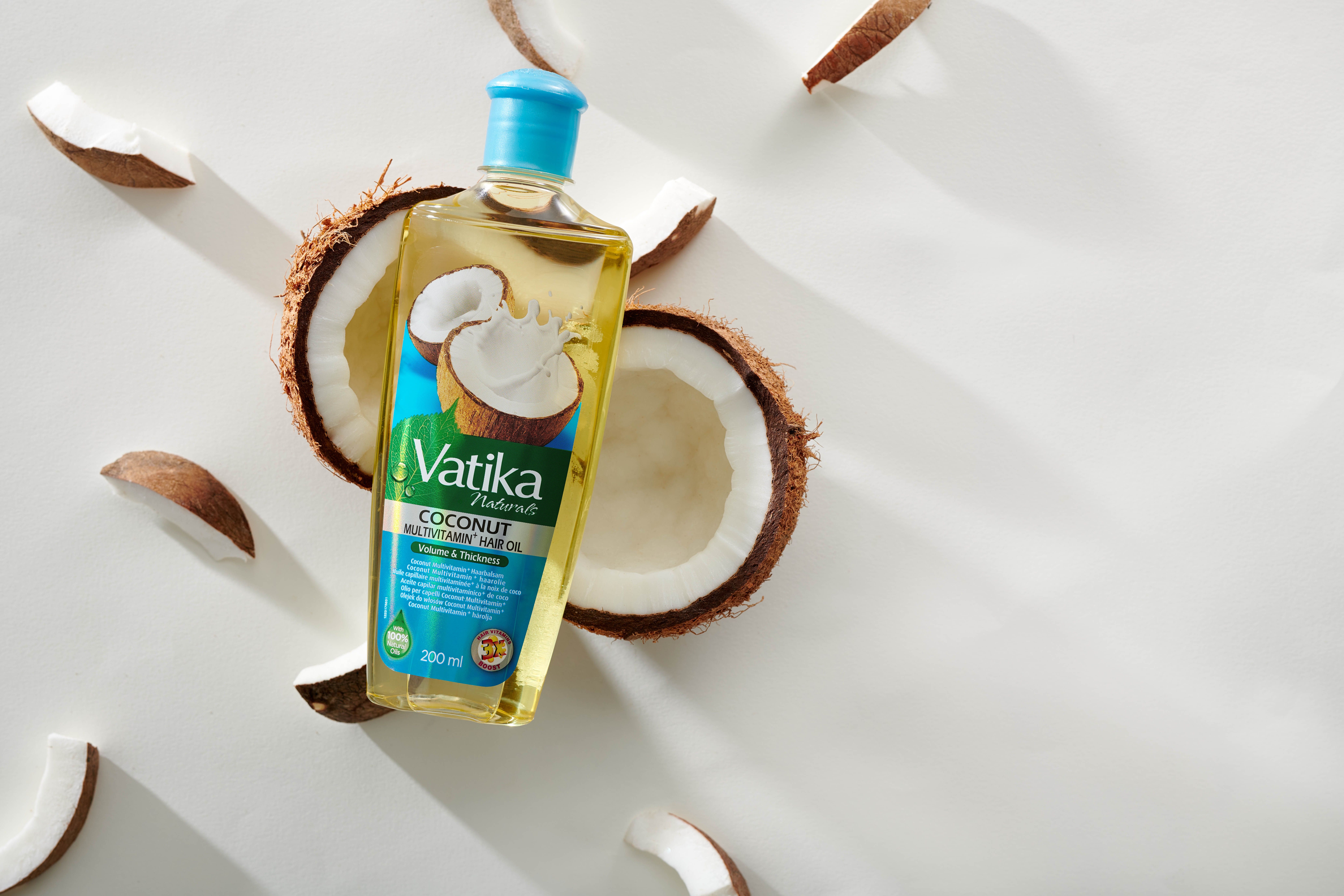 Vatika Naturals haaroliefles met blauwe dop, omringd door kokoshelften en -stukken. Opschrift: Coconut Multivitamin Hair Oil.