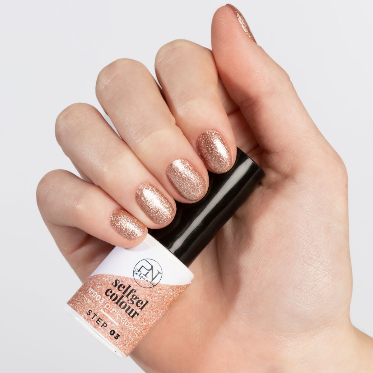 Main avec ongles vernis pailletés roses. Flacon de vernis à ongles avec bouchon noir. Inscription : PN Selfgel Colour.