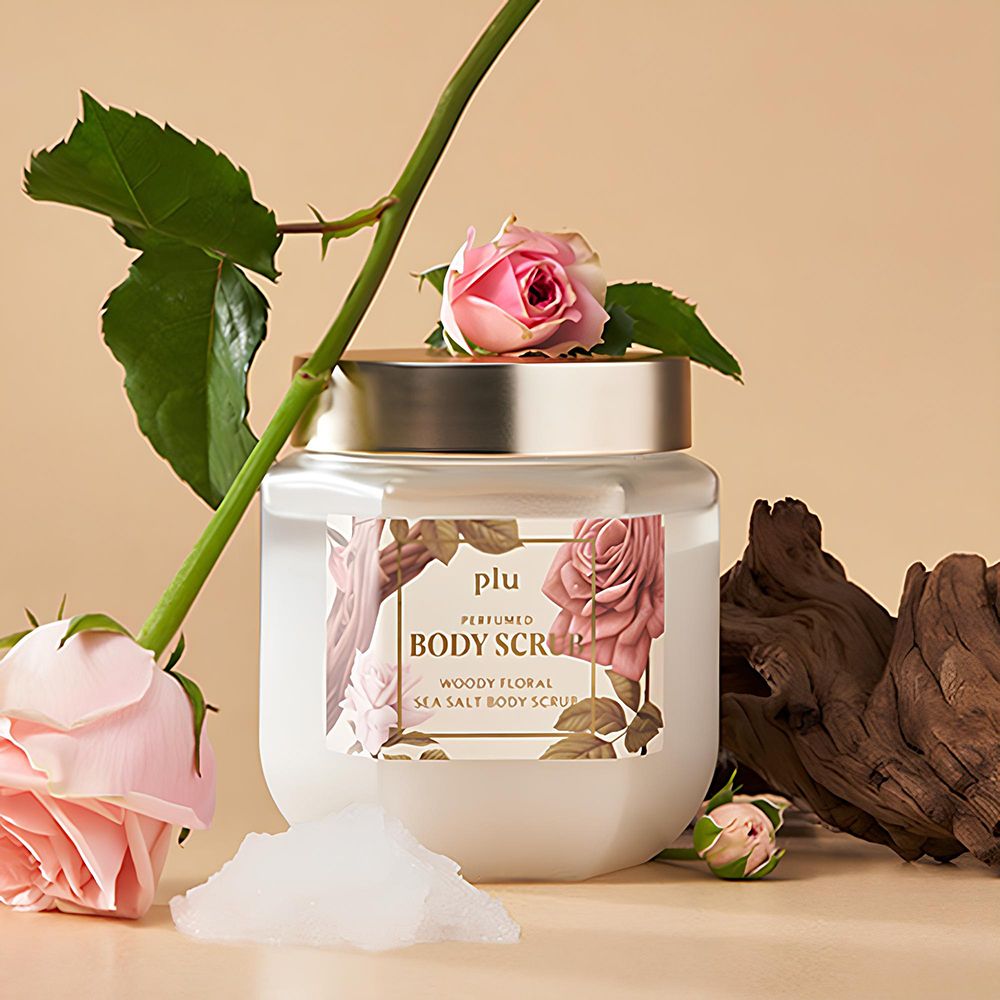 Pot en verre avec couvercle doré, entouré de roses et de bois. Étiquette avec nom du produit : PLU Perfumed Body Scrub.