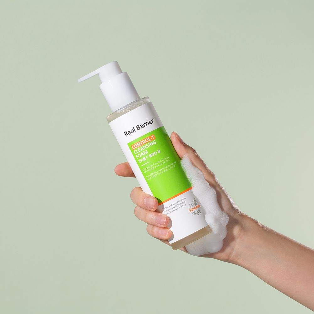 Wit-groene fles met pomp, vastgehouden door een hand. Schuim op de hand. Tekst: Real Barrier, Control-T Cleansing Foam.