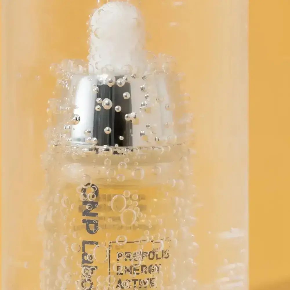 Close-up van een fles met geelachtige vloeistof en zilveren pipet. Bellen op de achtergrond. Opschrift: CNP Laboratory, PROPOLIS ENERGY ACTIVE AMPOULE.