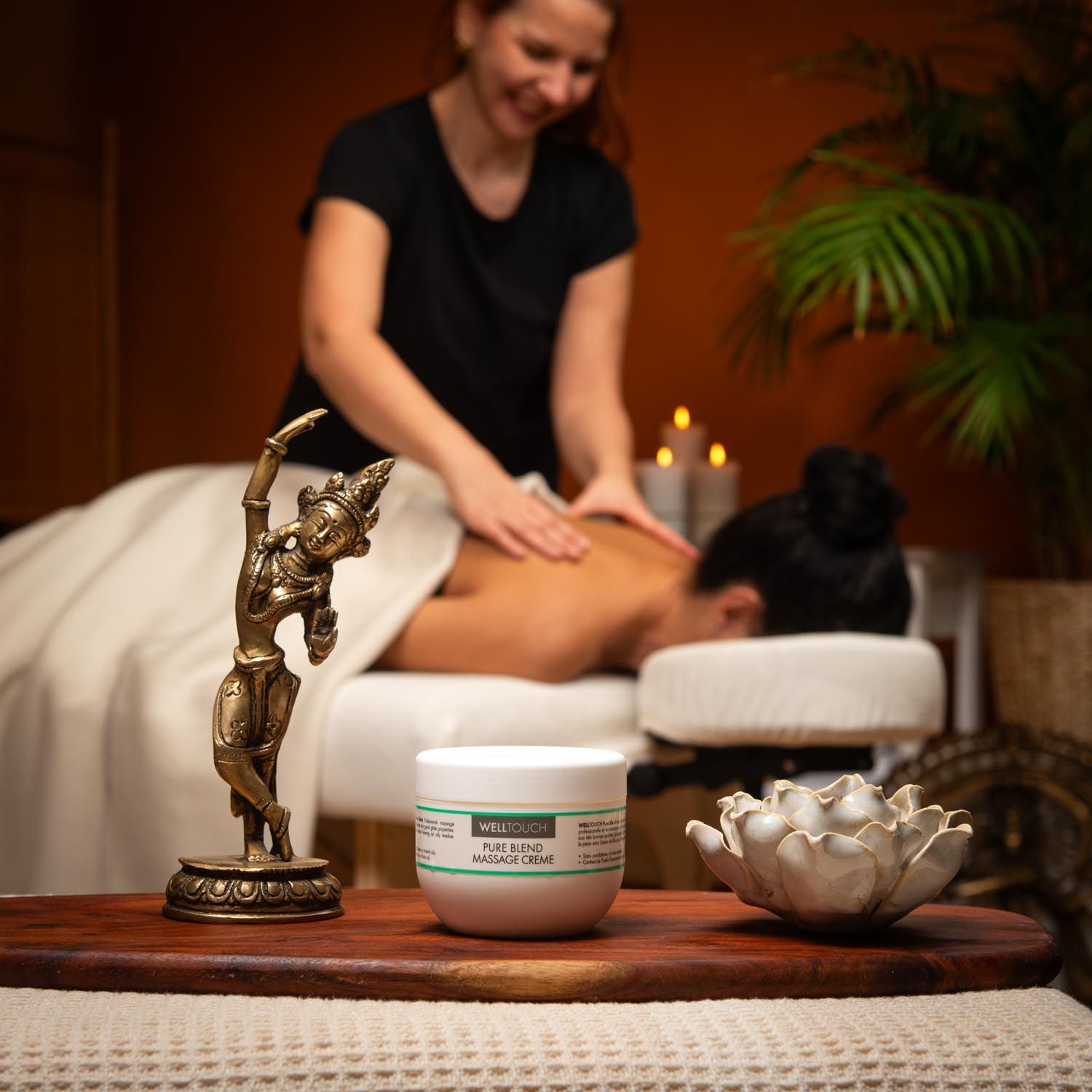 Witte crème pot met groene label. Opschrift: Welltouch Pure Blend Massage Creme. Op houten dienblad, voor massage scène.