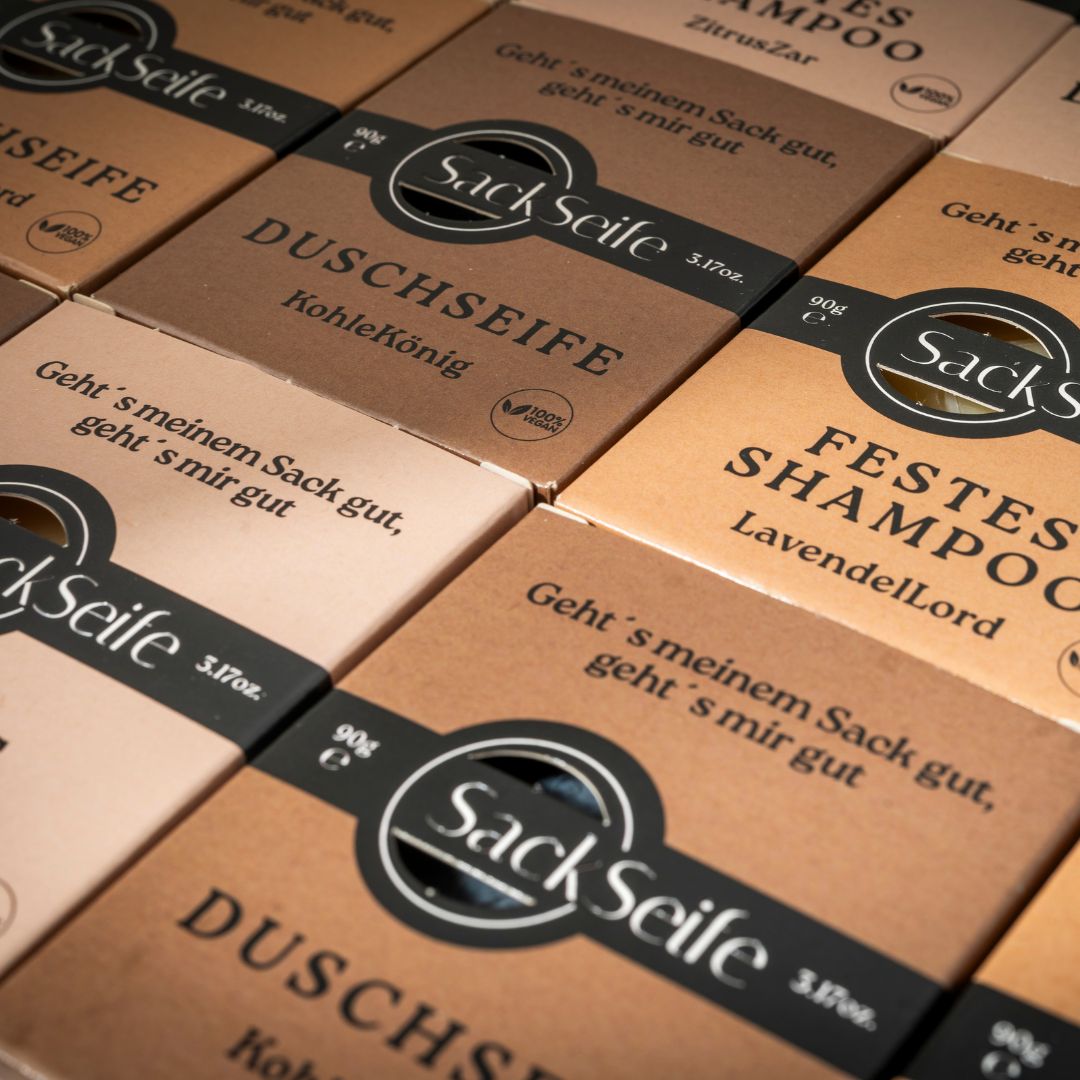 Gros plan de plusieurs boîtes. Inscriptions : SackSeife, DUSCHSEIFE, FESTES SHAMPOO. Emballage en carton brun.