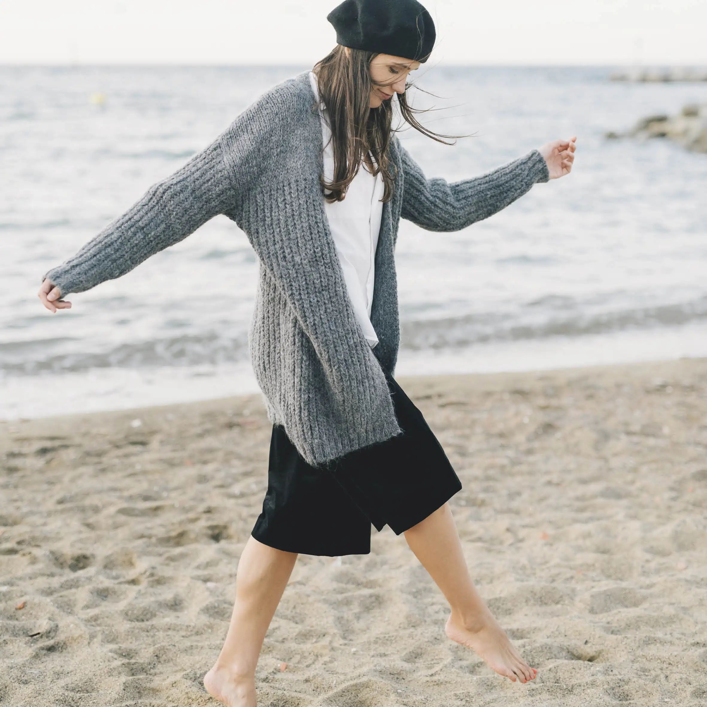 Femme en veste grise et béret noir, marchant sur la plage. Pieds nus, souriante.