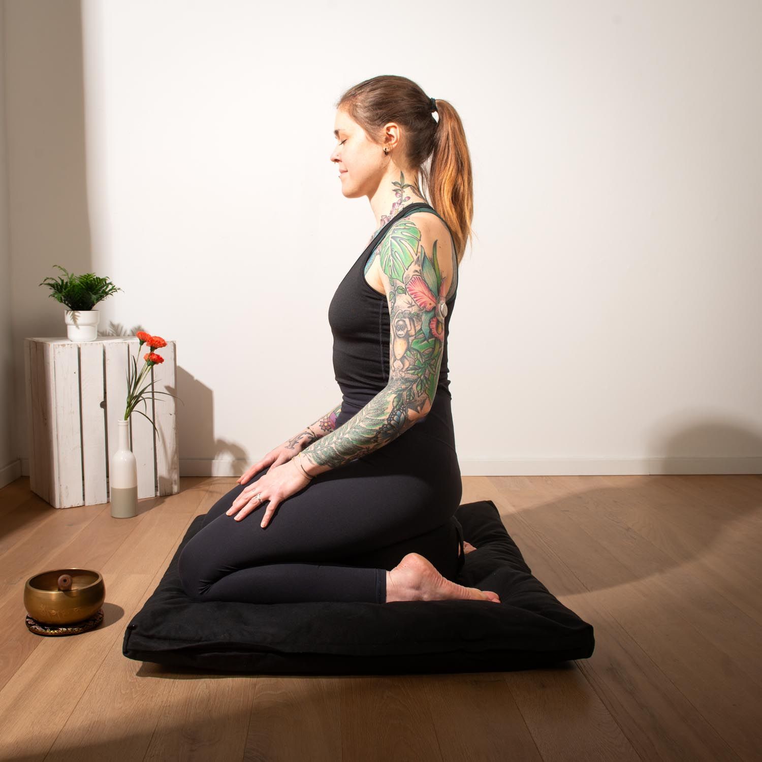 Personne assise sur un coussin et un tapis de méditation noirs. La personne porte des vêtements noirs.