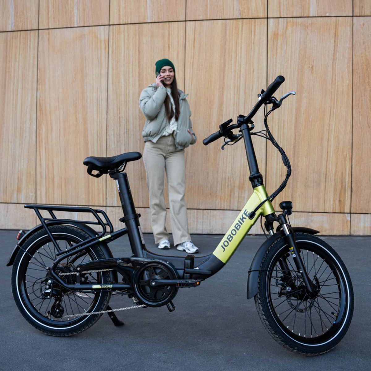 Vélo électrique pliable vert et noir. Logo JOBOBIKE. Porte-bagages, garde-boue. Personne téléphonant près du vélo.