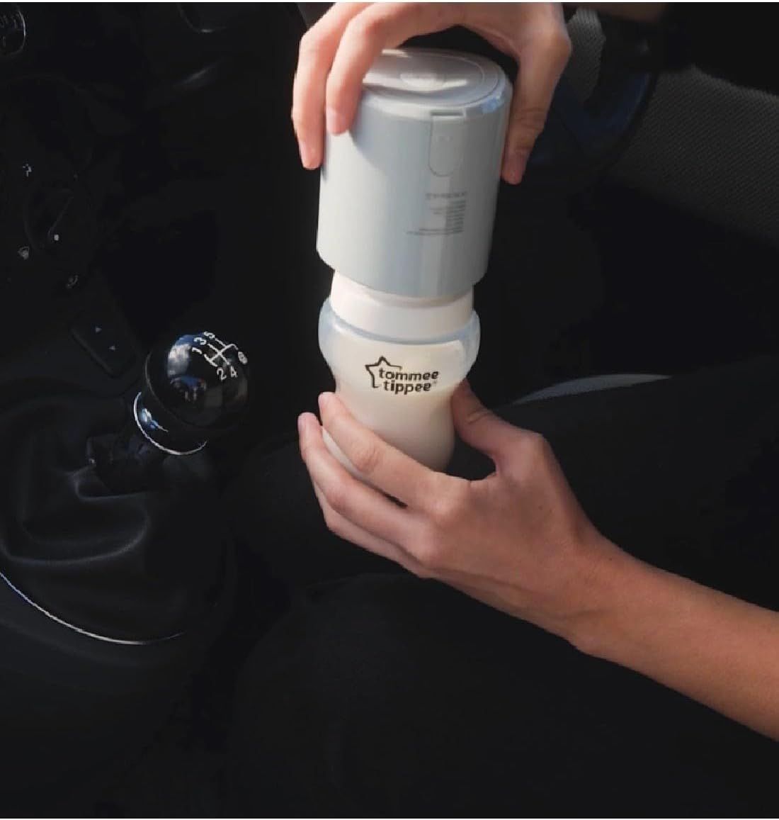 Draagbare flessenwarmer in lichtgrijs en wit, met babyfles. Handen houden het apparaat in de auto.