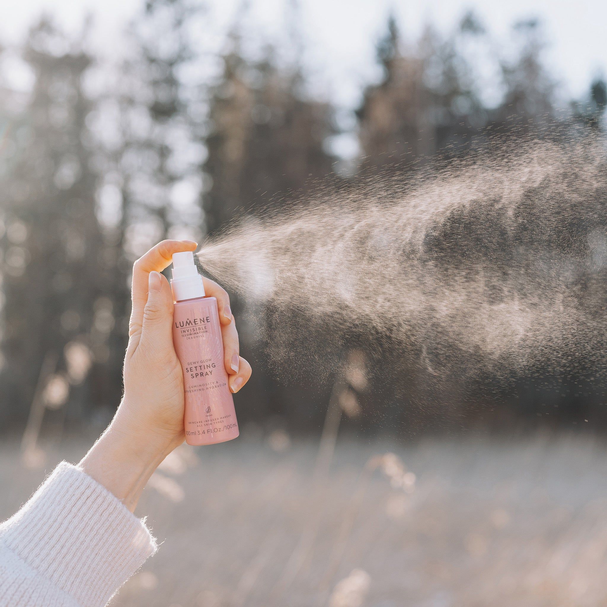 Hand houdt roze sprayfles vast. Nevel. Opschrift: Lumene INVISIBLE ILLUMINATION Dewy Glow Setting Spray. Achtergrond: natuur, bomen.