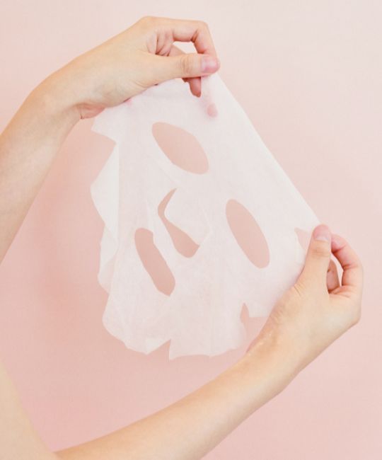 Masque facial blanc avec ouvertures pour les yeux, le nez et la bouche, tenu par deux mains sur fond rose.