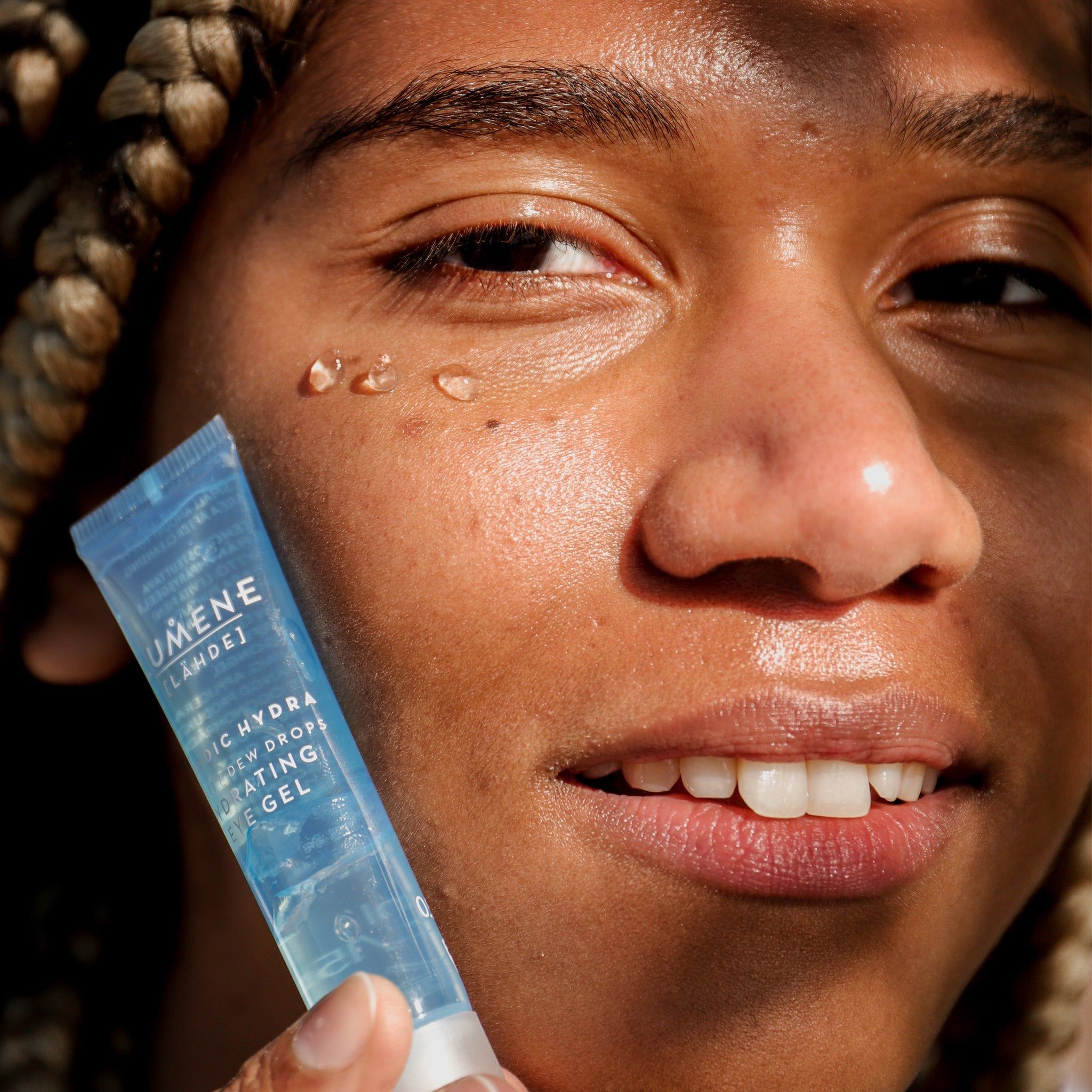 Personne tenant un tube devant son visage. Inscription : Lumene [LAHDE] Nordic Hydra Purity Dew Drops Hydrating Eye Gel. Gouttes de gel sur la peau.