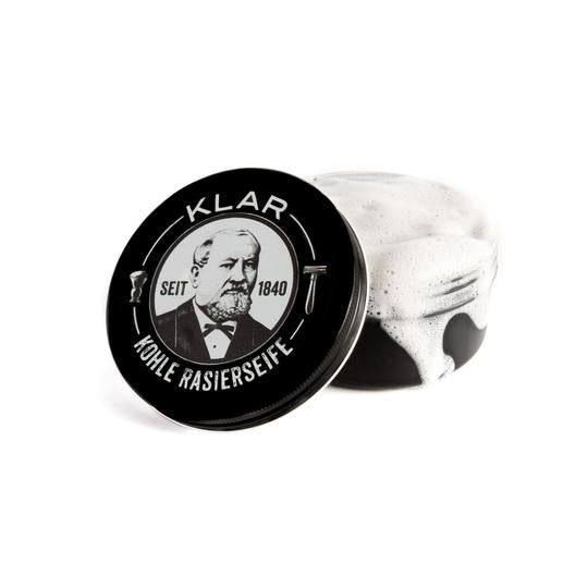 Boîte de savon à raser au charbon actif ouverte avec mousse. Couvercle avec logo et portrait. Inscription: KLAR, KOHLE RASIERSEIFE, SEIT 1840.