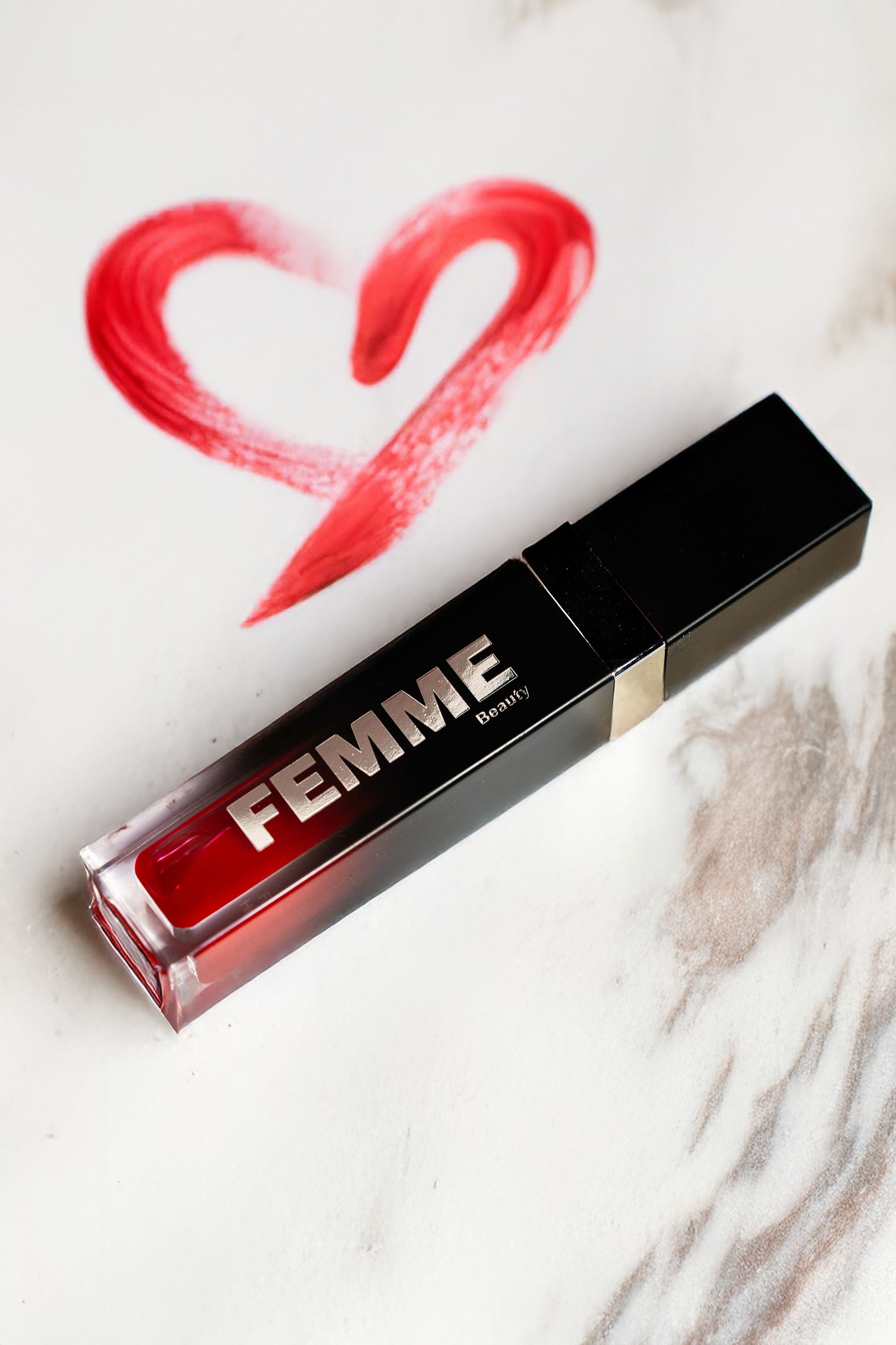 Huile à lèvres FEMME Beauty rouge, dans un tube transparent avec un capuchon noir. Un trait de pinceau rouge en forme de cœur à côté.