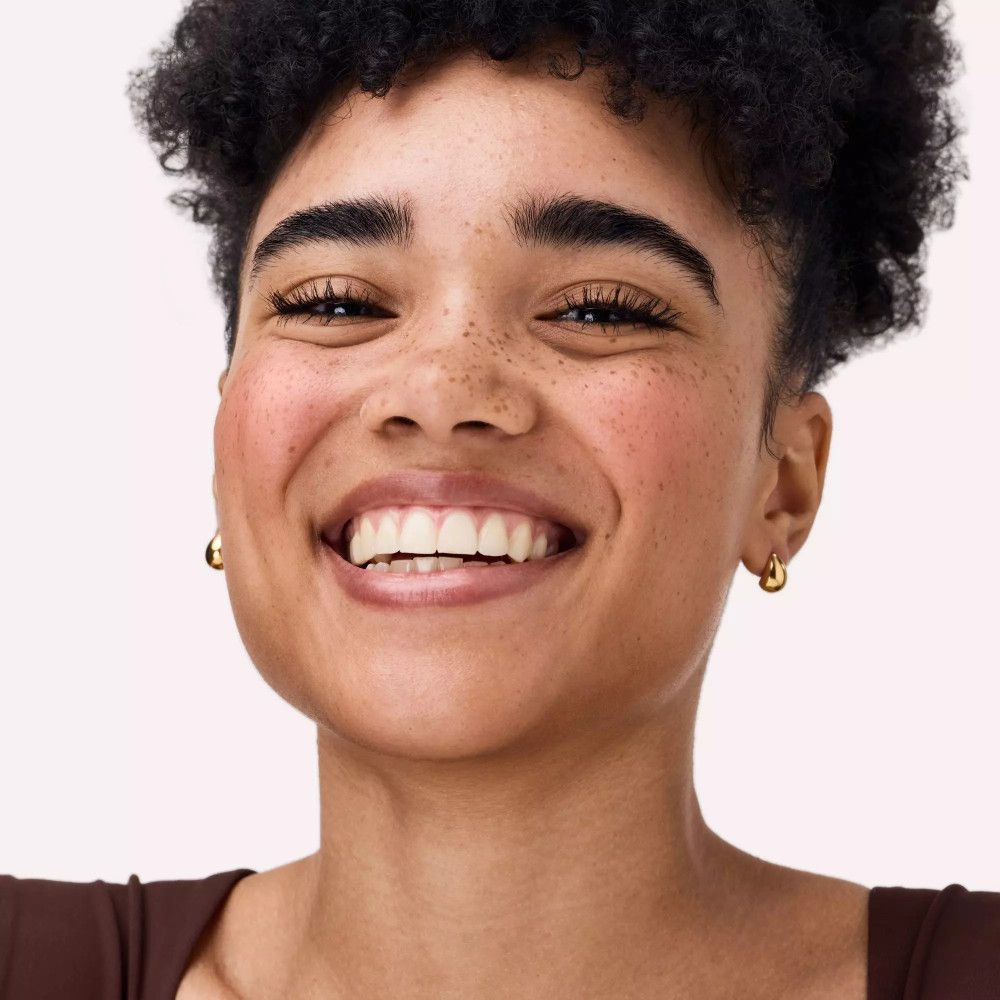 Gros plan d'une femme souriante avec du fard à joues sur les joues. Elle porte des boucles d'oreilles dorées.